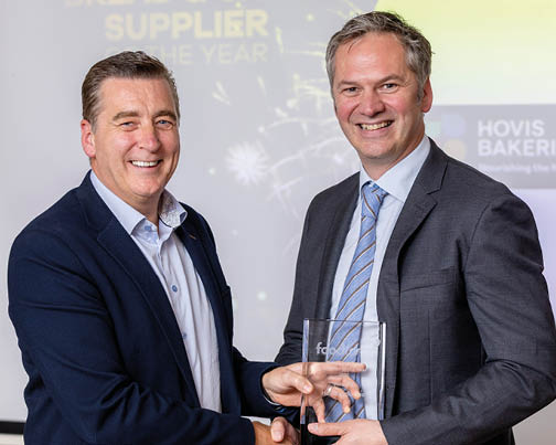 Two adults in business attire smile and shake hands while holding a clear award in a professional setting. Description generated by AI