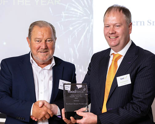 Two adults in business attire are smiling and shaking hands while holding an award plaque at a formal event. Description generated by AI