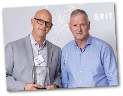 Two adults stand side by side, one wearing glasses and a light gray blazer holding a clear award, while the other wears a blue checkered shirt. Description generated by AI