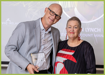 Two adults stand closely together smiling, with one holding a clear award and wearing glasses and a light gray blazer, while the other wears a dark top with a colorful bird design and red earrings. Description generated by AI