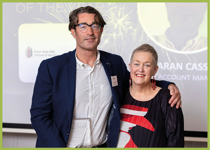 Two adults stand closely together smiling, one wearing glasses and a blazer with a name tag, and the other wearing a patterned dress with red earrings, in front of a presentation screen. Description generated by AI
