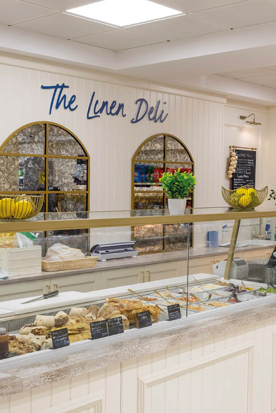 A bright deli counter with a glass display filled with various breads and pastries is set against a white paneled wall with decorative arched mirrors, potted plants, and a sign reading The Linen Deli. Description generated by AI