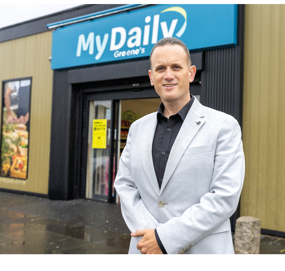A person wearing a light gray blazer and black shirt stands smiling outside a store with a sign partially visible above the entrance. Description generated by AI