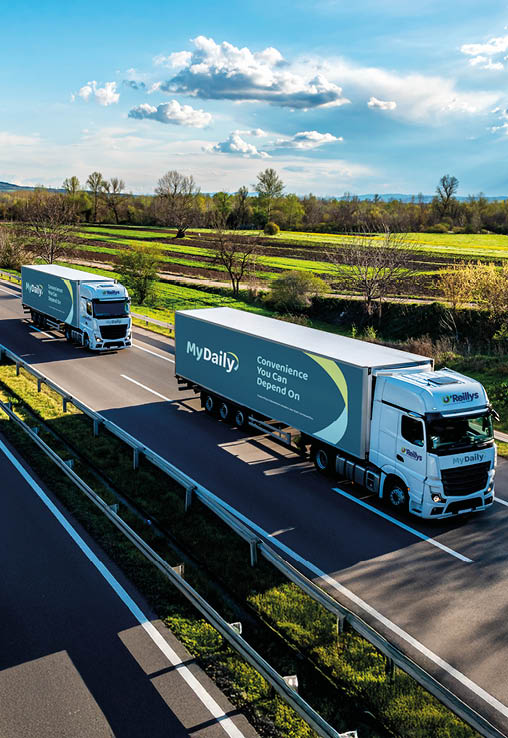 Three white trucks with trailers are driving in a line on a divided highway surrounded by green fields under a partly cloudy sky. Description generated by AI