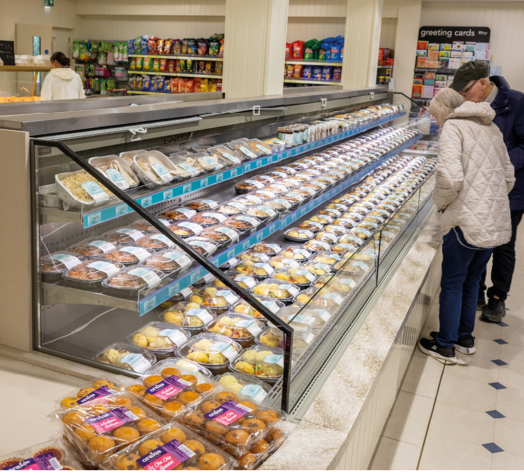 People in a store aisle are looking at a long refrigerated display case filled with packaged baked goods and snacks. Description generated by AI