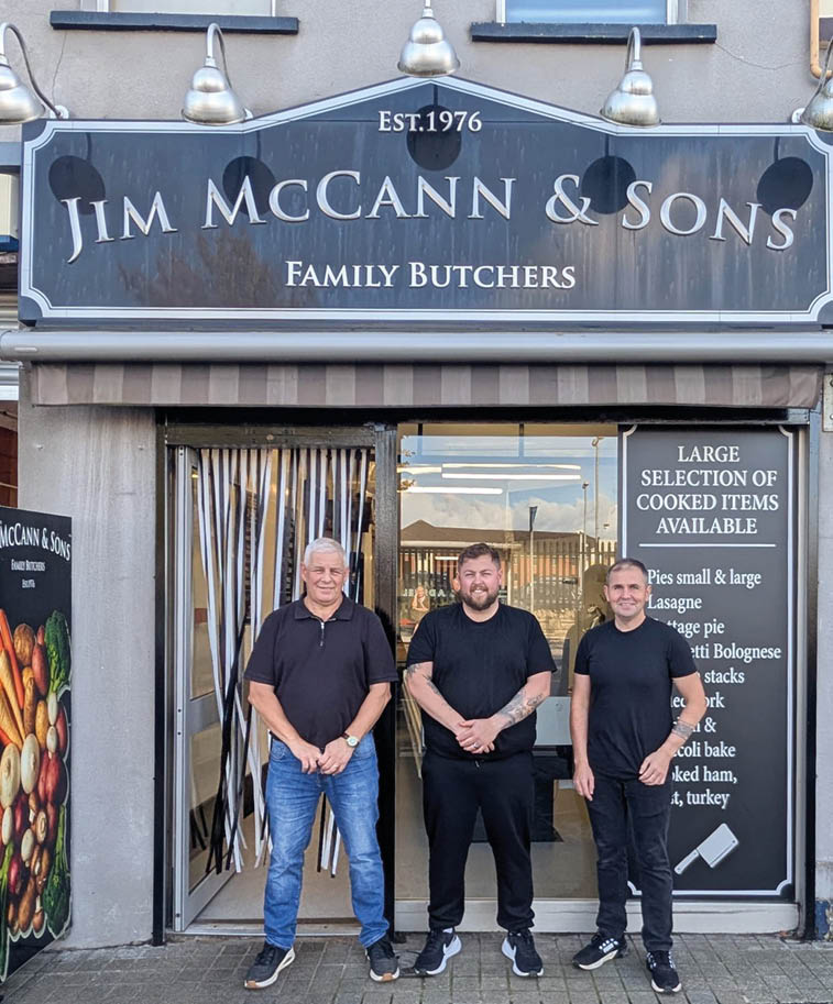 Three adults wearing casual black shirts and jeans stand smiling in front of a family butcher shop with a sign reading Jim McCann & Sons established 1976. Description generated by AI