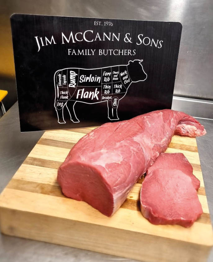 A wooden cutting board holds a large piece of raw beef with two slices cut from it, placed in front of a black sign for Jim McCann & Sons Family Butchers showing a diagram of beef cuts. Description generated by AI