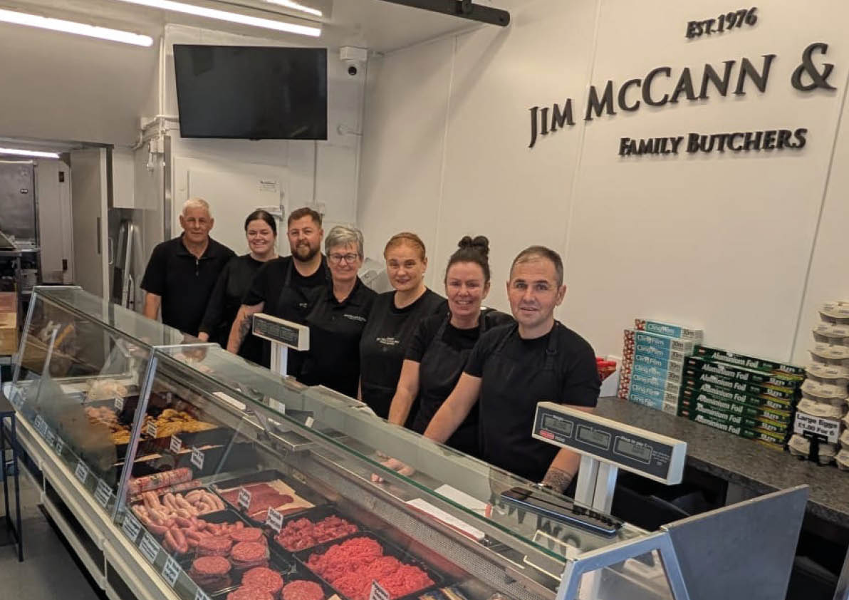 Seven adults wearing black shirts stand behind a glass display case filled with various cuts of meat inside a butcher shop with the sign Jim McCann & Sons Family Butchers on the wall. Description generated by AI