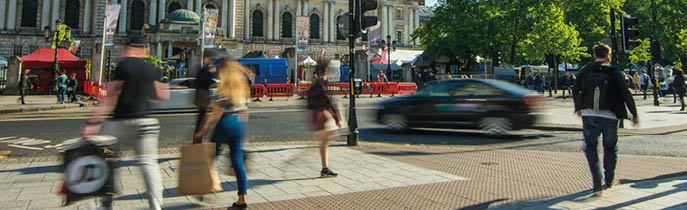 People walk and a car drives past a historic building with ornate architecture under a clear blue sky. Description generated by AI