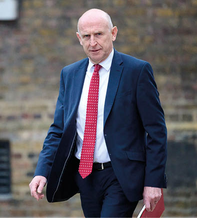 A person wearing a dark suit and red patterned tie is walking outdoors in front of a brick wall. Description generated by AI