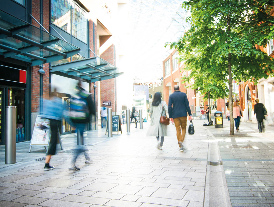 People walk along a bright, modern outdoor shopping street with brick buildings, trees, and glass canopies. Description generated by AI