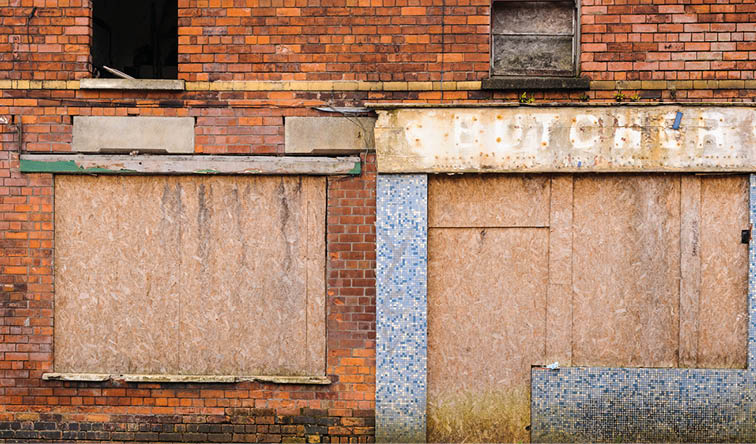 Two boarded-up windows on a brick wall, one framed with blue mosaic tiles and the other plain, showing signs of age and weathering. Description generated by AI