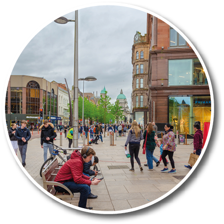 People walk and sit along a busy urban street lined with shops and historic buildings under a cloudy sky. Description generated by AI