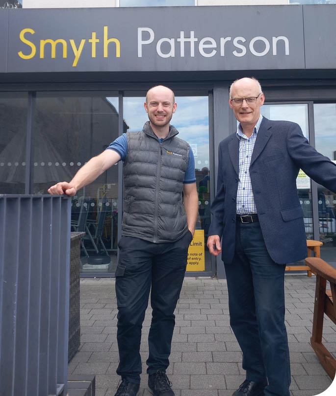 Two adults stand smiling outside a building with a sign that reads Smyth Patterson, one wearing a suit jacket and the other wearing a puffer vest. Description generated by AI