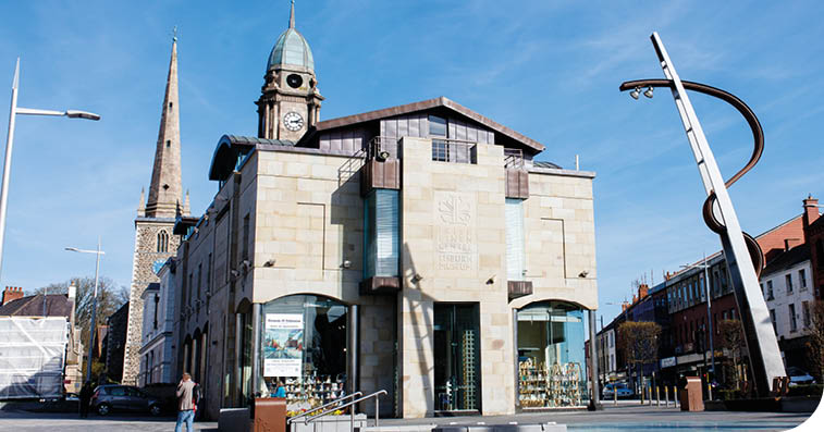 A historic stone building with large windows stands at a street corner under a clear blue sky, surrounded by street lamps and adjacent buildings. Description generated by AI