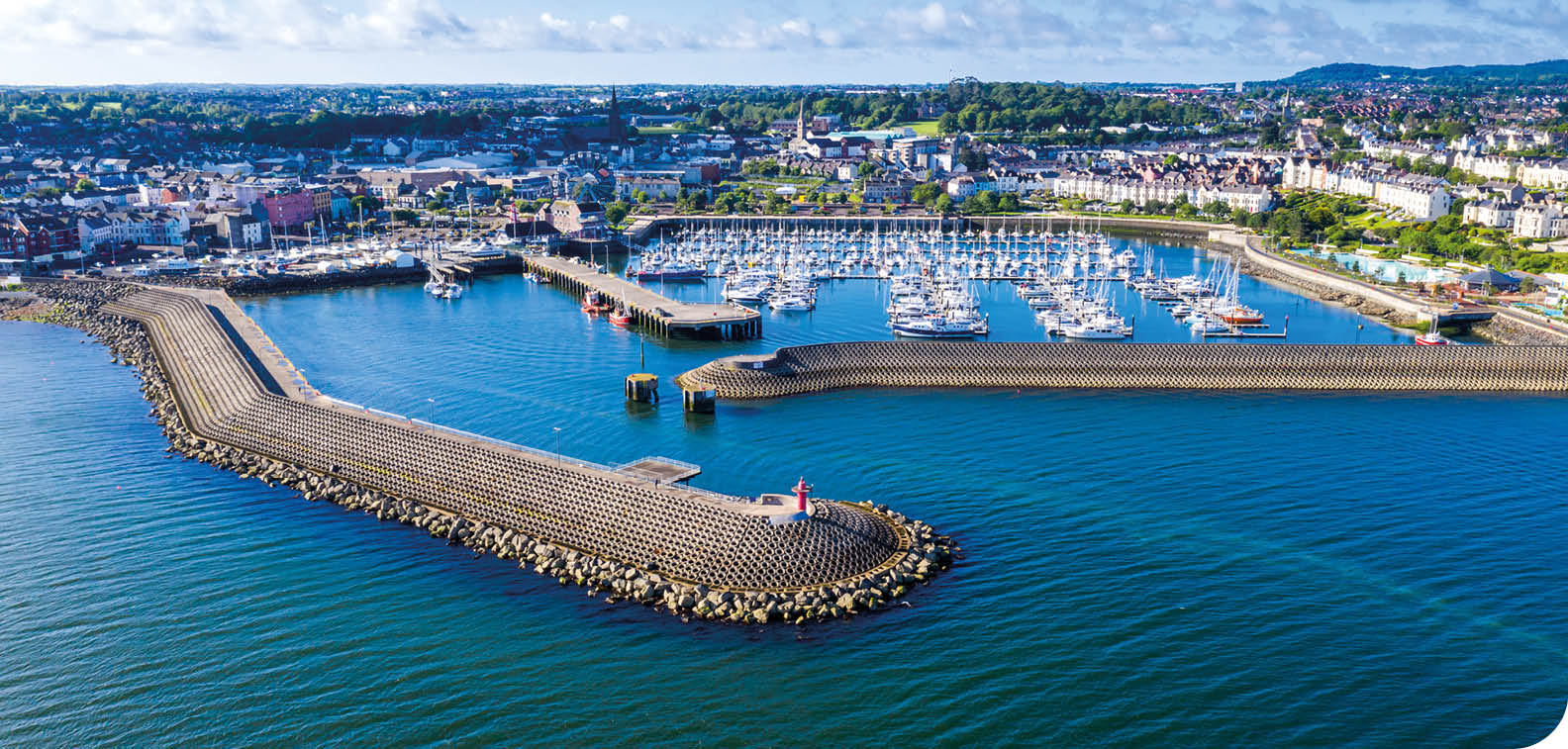 A coastal marina with numerous docked sailboats and yachts surrounded by a breakwater extending into calm blue water with a town and green hills in the background under a partly cloudy sky. Description generated by AI
