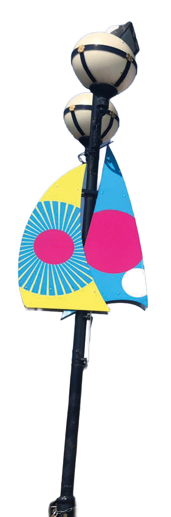 A tall black street lamp with two spherical lights has two colorful abstract flags attached, featuring bright yellow, blue, pink, and white shapes. Description generated by AI