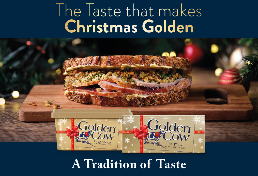 A sandwich with multigrain bread and filling is placed on a wooden board with two packages of Golden Cow butter and cassis spread in front, accompanied by festive Christmas decorations and the text promoting a golden Christmas taste tradition. Description generated by AI