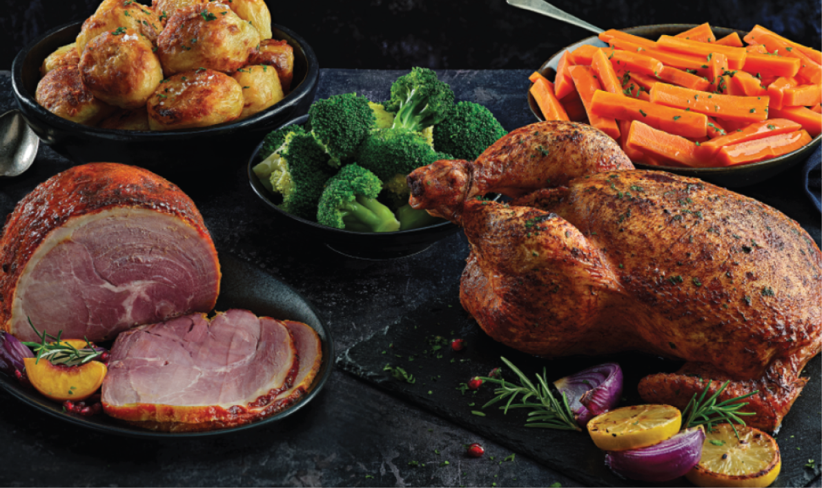 A roasted whole chicken is served alongside sliced ham, steamed broccoli, roasted potatoes, and glazed carrots on a dark surface. Description generated by AI