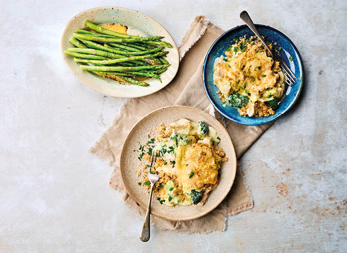 Two plates of baked casserole with broccoli and cheese are placed on a beige cloth napkin next to a plate of cooked asparagus on a light-colored surface. Description generated by AI