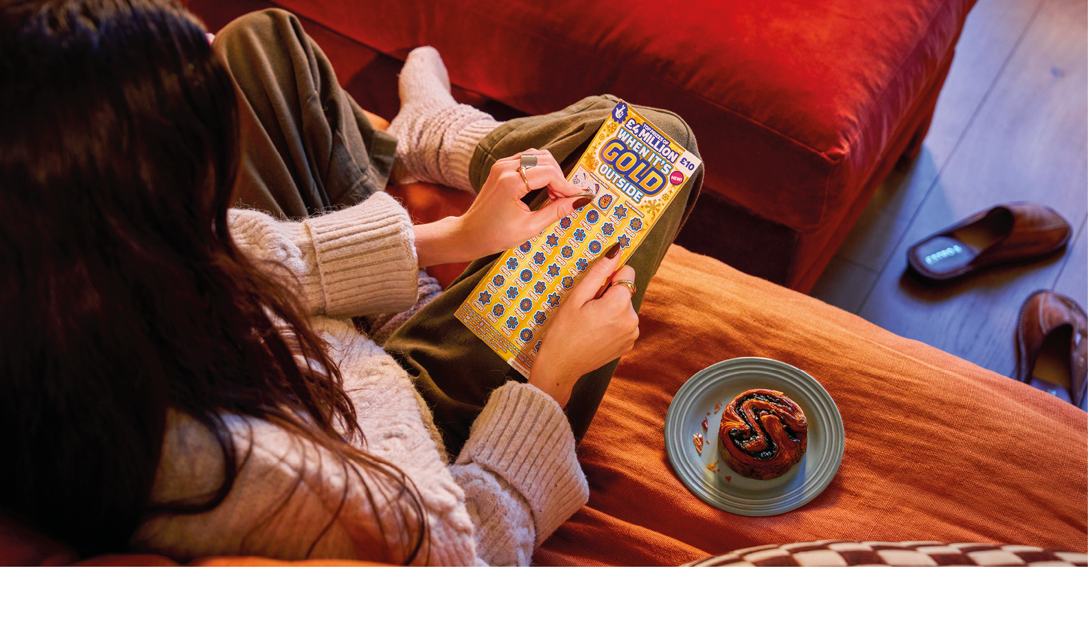 A person with long hair wearing a cozy sweater and pants is sitting on a couch holding a scratch-off lottery ticket while a small dessert on a plate rests nearby. Description generated by AI
