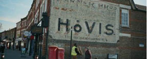 Two people walk past a brick building with a large faded advertisement for Hovis bread painted on the wall. Description generated by AI