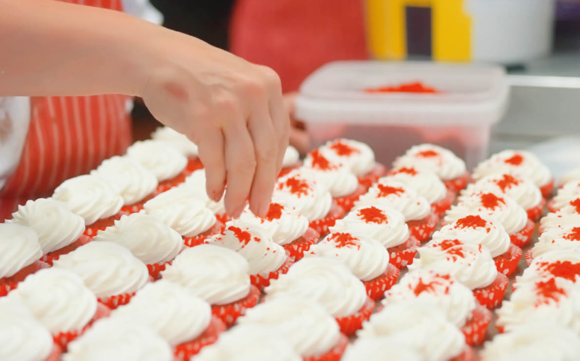 A person wearing a striped apron is carefully placing red sprinkles on rows of white frosted cupcakes in a kitchen setting. Description generated by AI