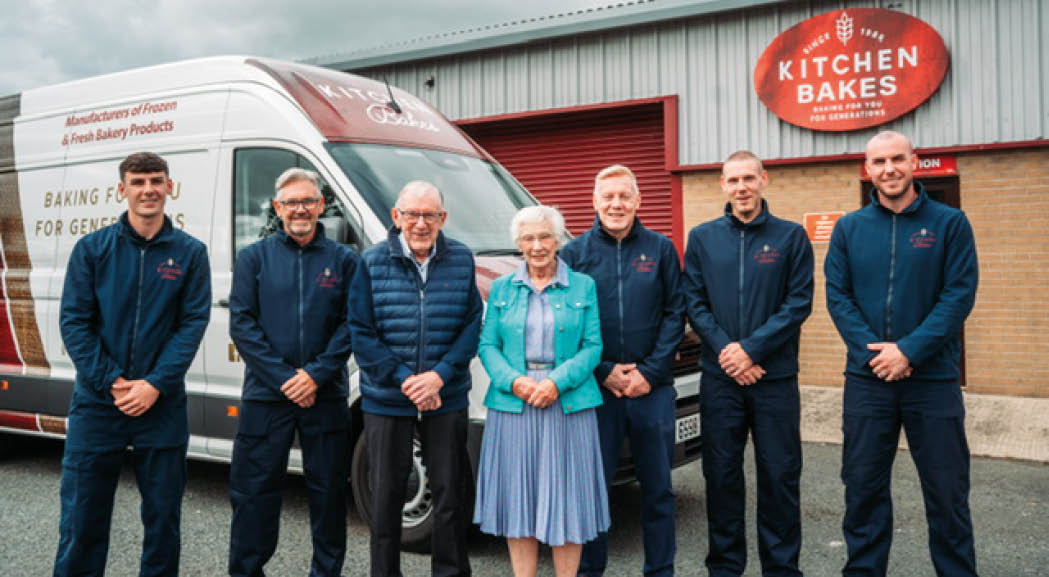 Seven adults wearing matching dark jackets and one adult in a blue dress and turquoise jacket stand smiling in front of a bakery van and building with a Kitchen Bakes sign. Description generated by AI