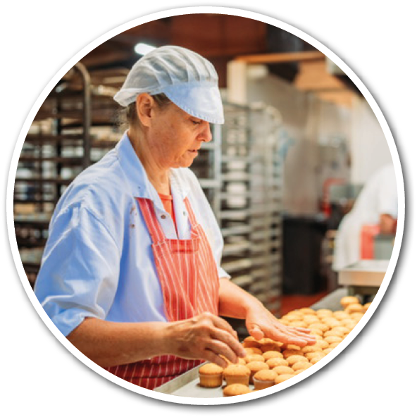 A person wearing a striped apron is arranging rows of small baked goods on a tray in a bakery setting. Description generated by AI