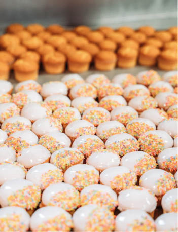 Rows of small round pastries are arranged on trays, with the front ones topped with white icing and colorful sprinkles and the back ones plain and golden brown. Description generated by AI