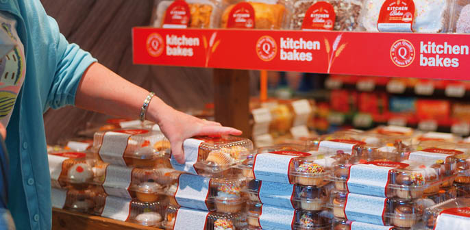 A person wearing a blue shirt is reaching to pick up a packaged baked good from a display table filled with stacks of similar packaged baked goods in a store. Description generated by AI