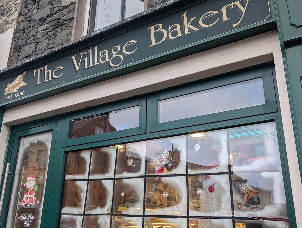 The Village Bakery storefront features a green frame with frosted windows decorated with festive holiday stickers and displays inside. Description generated by AI