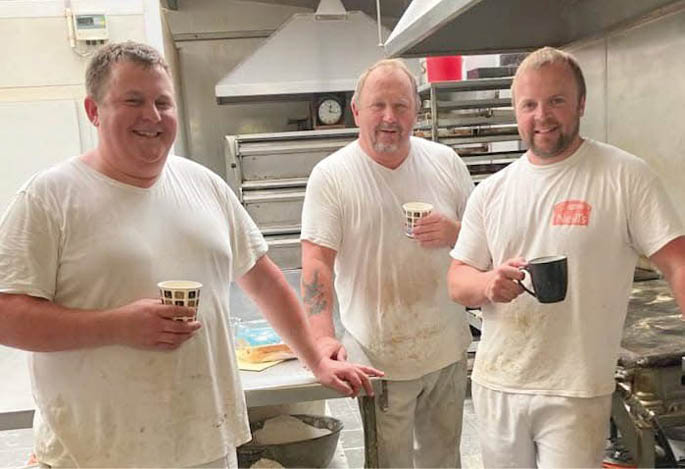 Three adults wearing white work clothes are standing in a kitchen or bakery setting, each holding a cup and smiling. Description generated by AI