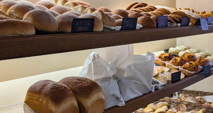 Shelves filled with various types of bread and baked goods are displayed in a well-lit bakery setting. Description generated by AI