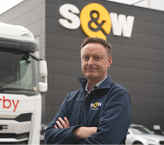 A person wearing a navy jacket with a company logo stands with arms crossed in front of a white truck and a building displaying the same company logo. Description generated by AI