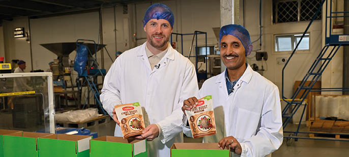 Two people wearing white lab coats and blue hairnets are standing in a factory setting holding packaged food products above green boxes on a conveyor belt. Description generated by AI