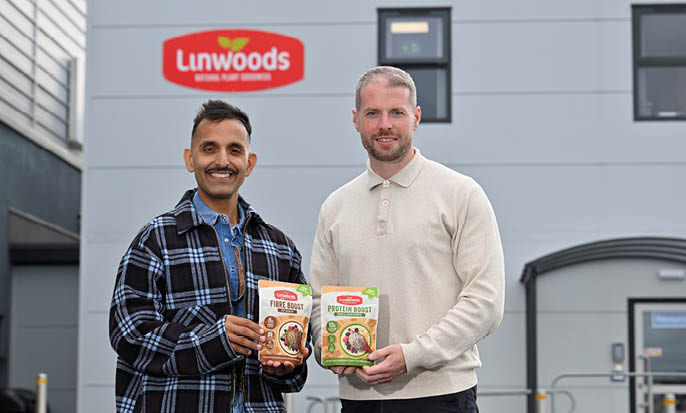 Two adults stand outside a building with a Linwoods sign, smiling and holding packaged food products. Description generated by AI