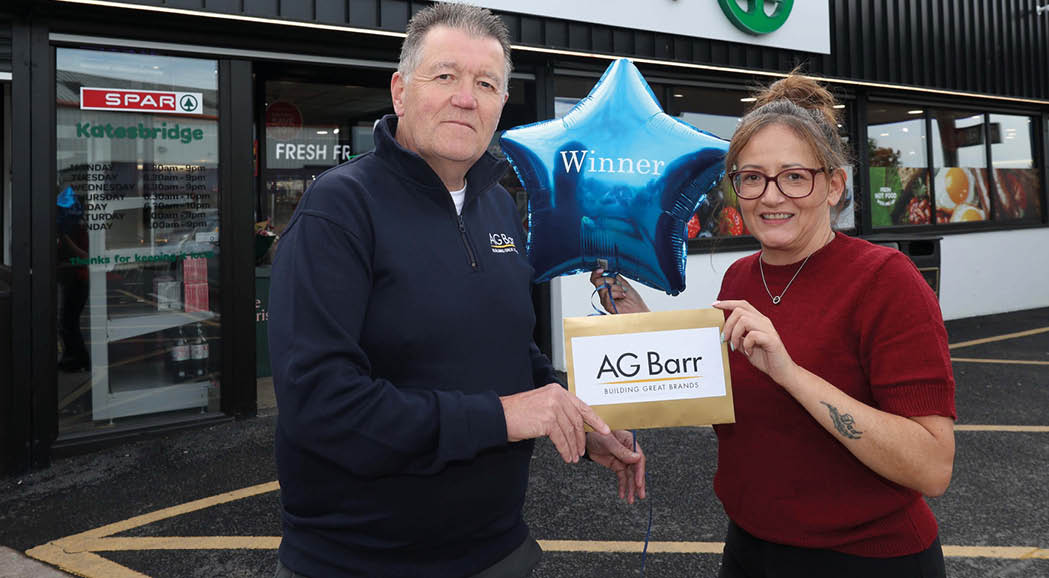 Two adults stand outside a store holding a blue star-shaped balloon that says “Winner" and an envelope labeled "AG Barr." Description generated by AI