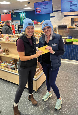 Two adults wearing matching blue beanies and black vests smile and hold a yellow envelope inside a grocery store near a bakery section. Description generated by AI