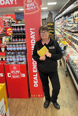 A person wearing glasses and a black jacket stands in a grocery store aisle holding a yellow envelope near a display of promotional signs advertising a deal ending Sunday. Description generated by AI