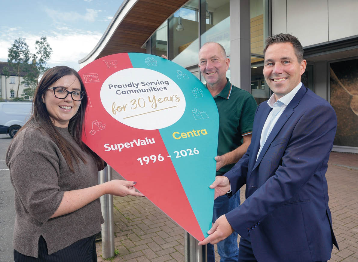 Three adults smiling and holding a large colorful sign shaped like a location pin that celebrates 30 years of service by SuperValu and Centra outside a modern building. Description generated by AI