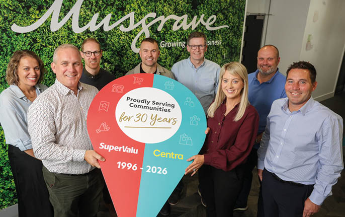 A group of eight adults standing indoors in front of a green leafy wall with the word “Musgrave" holding a large colorful sign celebrating 30 years of community service by SuperValu and Centra. Description generated by AI