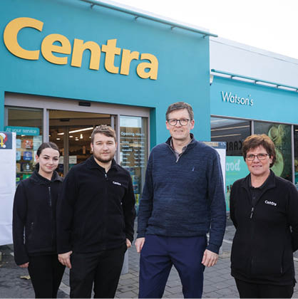 Four adults wearing black jackets stand outside a Centra store with a Watson's store visible in the background. Description generated by AI