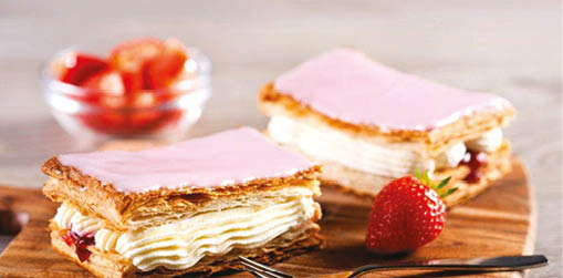 Two layered pastries with pink icing and cream filling are placed on a wooden board next to a strawberry and a fork. Description generated by AI