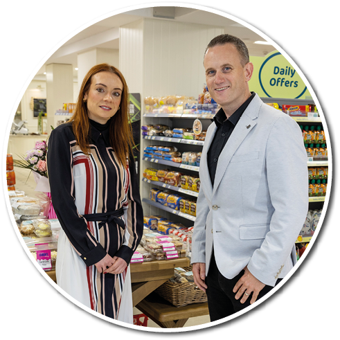 Two adults stand in a grocery store aisle near a wooden table displaying packaged bread and baked goods, with one wearing a striped dress and the other wearing a light-colored blazer and dark pants. Description generated by AI