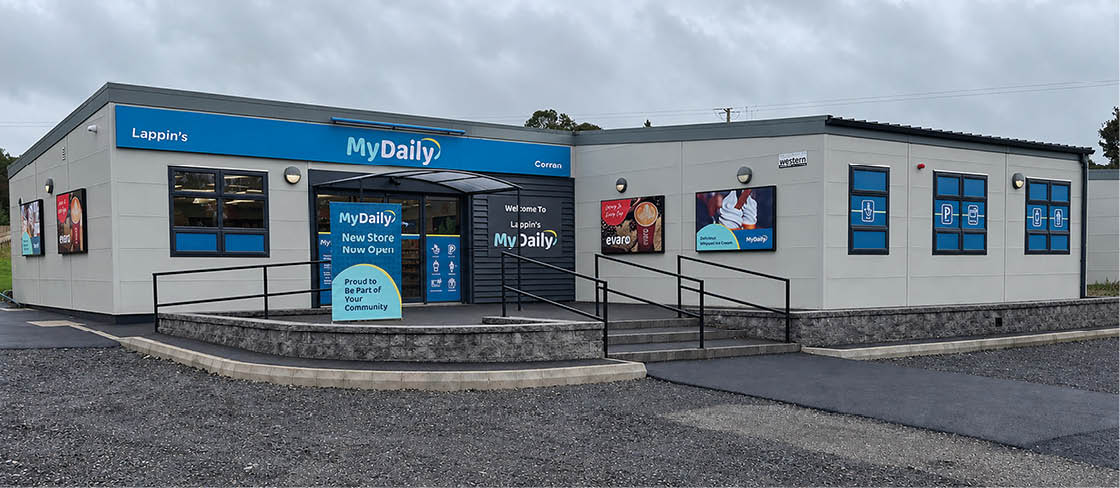 A storefront with a blue awning labeled MyDaily displays signs announcing a new store opening and community involvement, with accessible ramps and windows featuring promotional posters. Description generated by AI