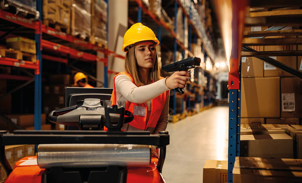 Female warehouse worker with safety helmet is working with bar code scanner while sitting on forklift in warehouse