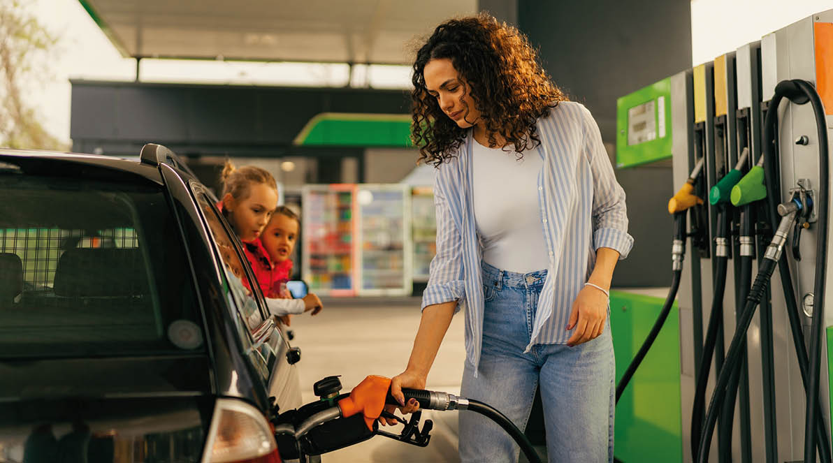 A young mother fills up gas tank at a gas station while her daughters look out the car window