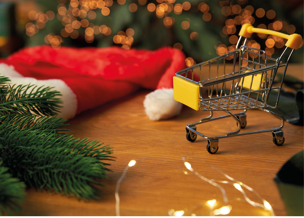Santa hat, Christmas tree branch and a small grocery cart on wooden background. Christmas and New Year background or backdrop. Side view