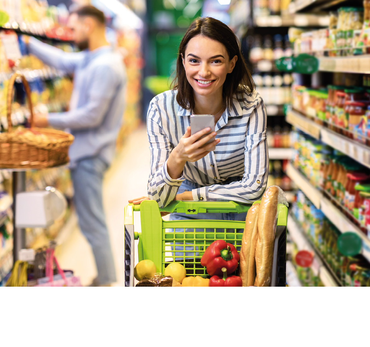 Portrait of smiling young casual woman leaning on shopping trolley cart indoors, using her mobile phone, posing and looking at camera. Satisfied female customer buying products in local grocery store